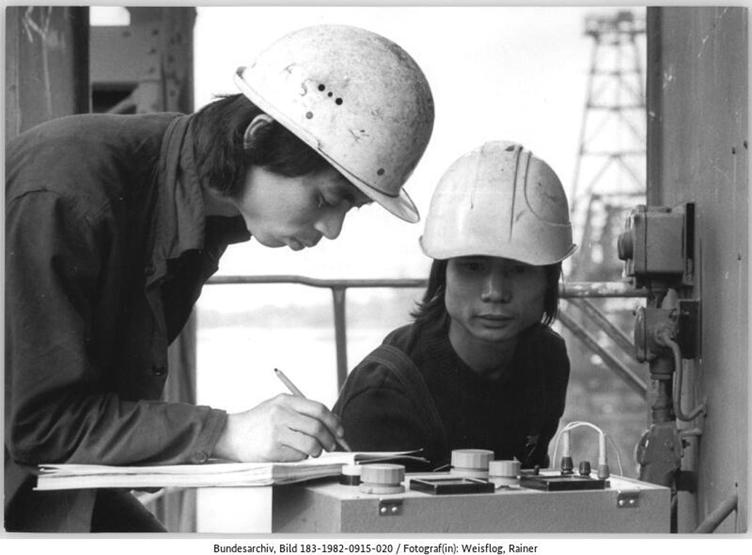 Zwei Männer in Arbeitskleidung und Schutzhelm beugen sich über eine technische Anlage. Einer der beiden, der links im Bild zu sehen ist, notiert etwas in ein Notizbuch.