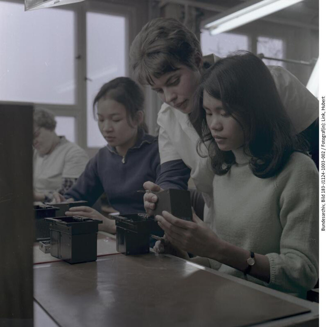 Nahaufnahme einer Werkbank im Betrieb, an der links und rechts zwei vietnamesische Vertragsarbeiterinnen sitzen. Zwischen ihnen beugt sich die deutsche Ausbilderin über die Schulter der rechtssitzenden Vertragsarbeiterin.