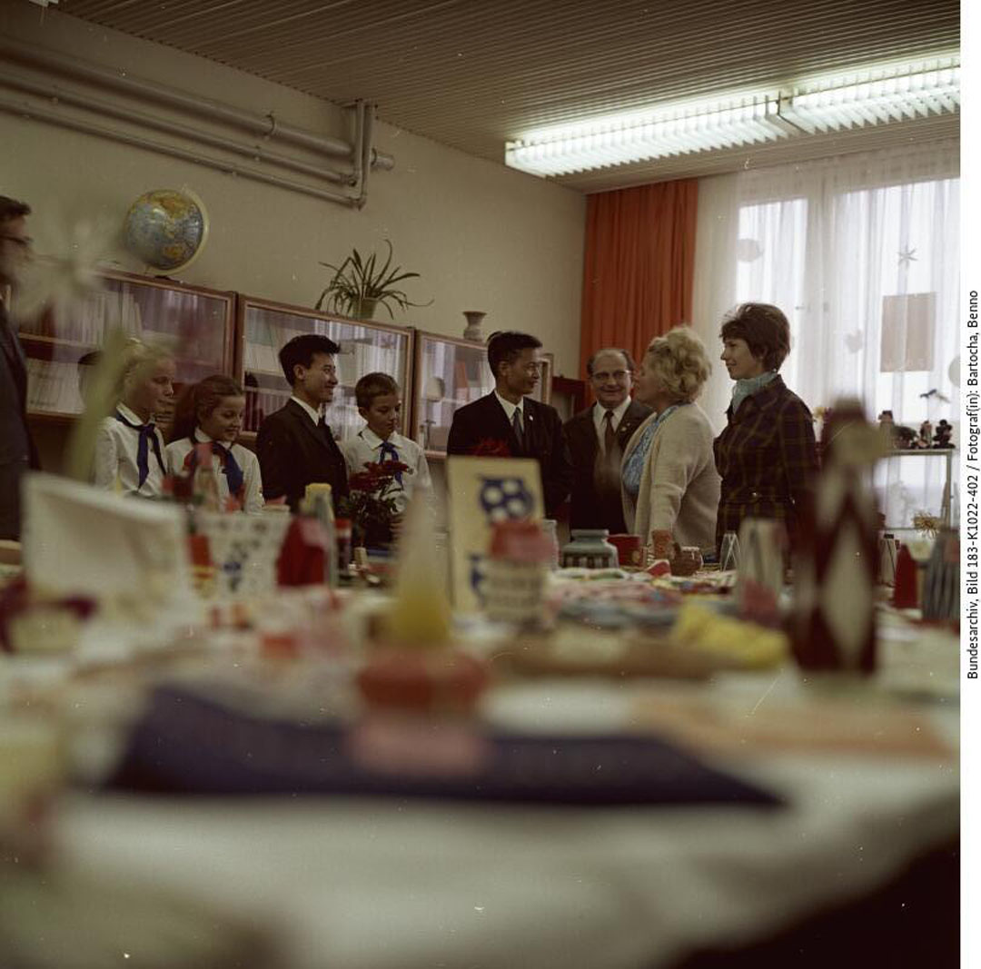 Blick in einen Klassenraum. Im Vordergrund sind Basteleien auf einem Tisch ausgestellt, die von Schulkindern hergestellt wurden. Im Hintergrund befindet sich eine Gruppe Menschen im Gespräch, darunter Pioniere und nordvietnamesische Botschaftsangehörige.