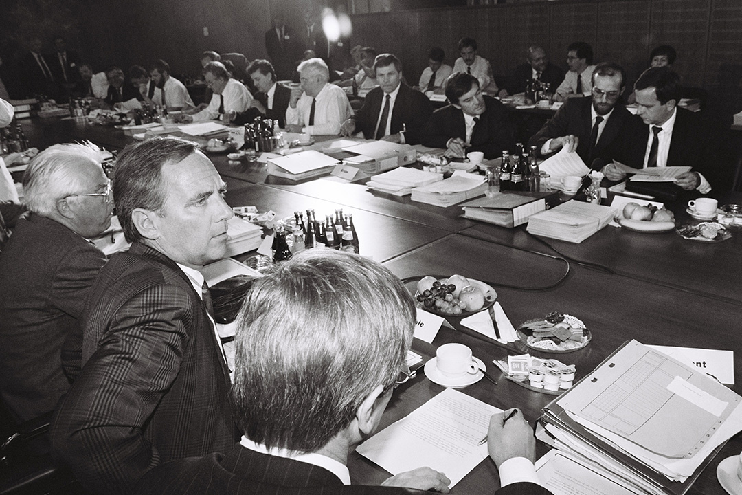 Zahlreiche Männer sitzen mit Dokumenten an einem langen Konerenztisch und scheinen über die Dokumente zu diskutieren. Auf dem Tisch stehen Kaffeetassen, Getränke und Obst.
