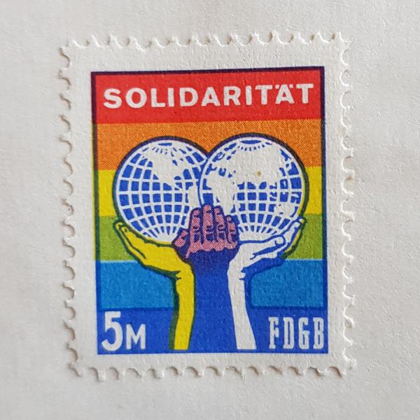 Solidaritätsmarke des FDGB im Wert von 5 Mark. Vor einem mehrfarbigen Hintergrund und unter der Überschrift „Solidarität“ halten drei Arme in gelber, brauner und weißer Farbe eine aufgeklappte Weltkugel. 