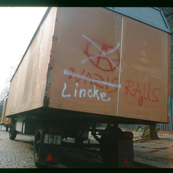 Zu sehen ist eine Farbfotographie eines braunen LKW-Anhängers, der am Straßenrad steht. Hinten hat jemand mit roter Farbe „NAZIS RAUS“ und das Antifa-Symbol „A“ in einem Kreis darauf geschrieben. Mit weißer Farbe wurde das Symbol und das Wort „NAZIS“ durchgestrichen und „Lincke“ daruntergeschrieben.