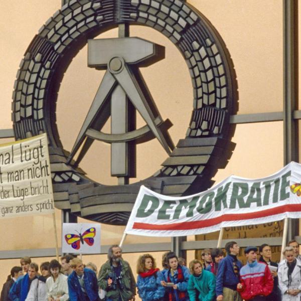 Menschen demonstrieren für Demokratie mit Bannern. Hinter Ihnen das Symbol der DDR 