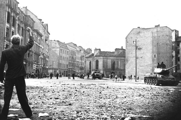 Eine Person steht mit erhobener Faust auf einer zerstörten Straße. Im Hintergrund sieht man einen Panzer