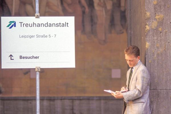 Ein Mann mit Aktenkoffer steht mit einem Buch in der Hand vor einem Schild, das auf den Besuchereingang der Treuhandanstalt verweist.